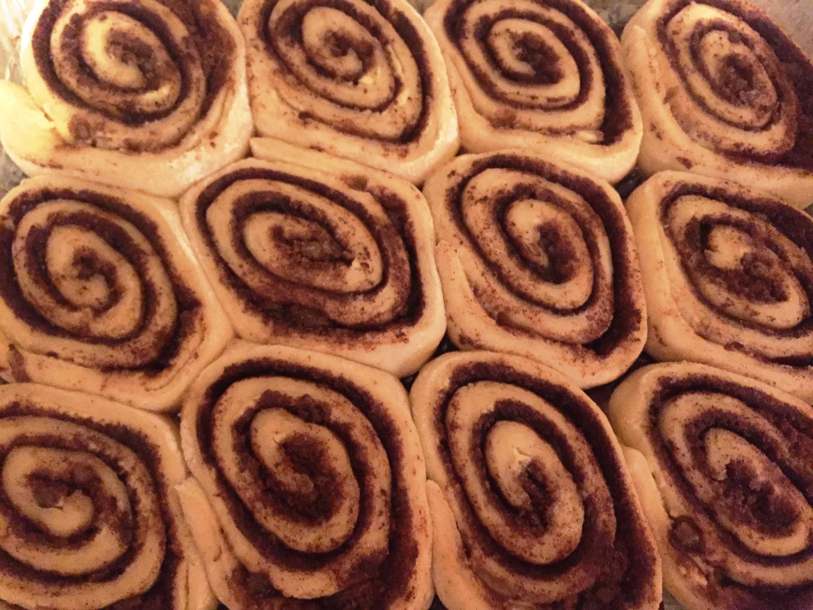 A close-up view of several uncooked cinnamon rolls arranged neatly in a baking tray. The cinnamon swirls are prominently visible, and the dough has a light yellow color. The rolls are tightly packed together, ready to be baked.